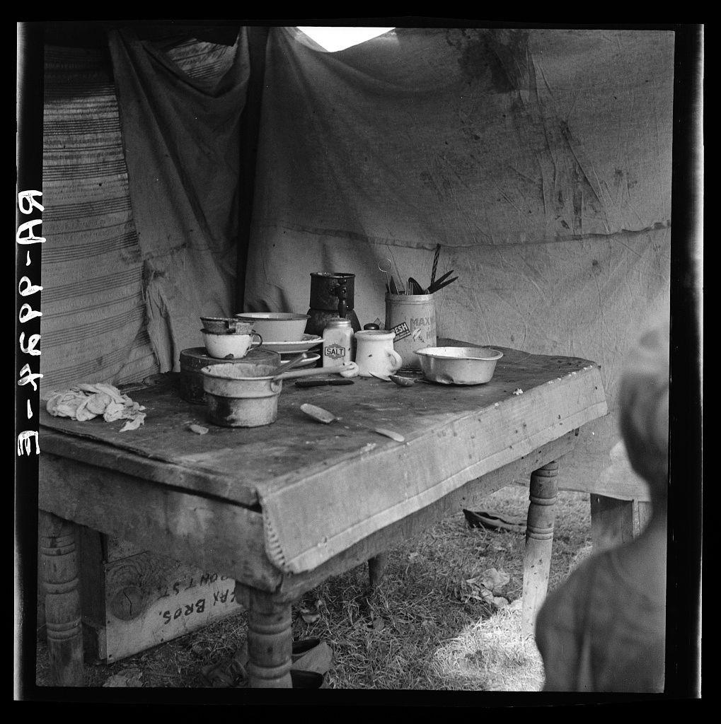 The image depicts a rustic kitchen setup in what appears to be an outdoor or makeshift shelter. A weathered wooden table is set with various cooking utensils and ingredients, such as bowls, cups, and a canister of salt labeled "SALT." There's also a container for 'FRESH MAX' food supplies visible. The setting suggests the living conditions might be challenging or temporary, possibly reflecting on-the-road life that migrant families often face during certain historical periods in America.

The photograph is monochromatic with clear contrasts and shadows playing across the scene, adding to its stark realism. Text overlayed in a vertical format at the top of the image appears to include letters such as 'A9' which could be an identification or cataloging code for this particular photo within a larger collection. The source indicates that the photograph is part of the "Food supply of migrant family" series by Dorothea Lange, documenting American River camp near Sacramento, California.