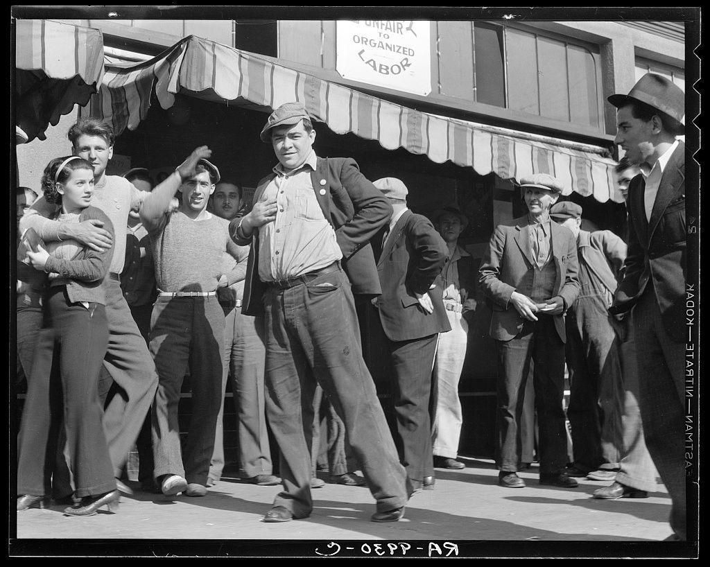 The image is a black and white photograph depicting a lively street scene. In the foreground, there's a man wearing a hat and jacket with rolled-up sleeves energetically dancing or gesturing in mid-motion on a sidewalk. He appears to be engaging directly with the camera, exuding confidence as he sticks out his chest. Behind him, various people are present: some appear engaged with each other while others focus on their surroundings.

The crowd includes men and women dressed in attire typical of an earlier era—men's suits, ties, hats; women wearing dresses or skirts and blouses, often accessorized with headbands or scarves. The expressions range from smiles to neutral looks as they observe the dancing man.

In the background, we can see a building structure that has signs indicating it might be related to labor unions: "UNFAIR TO ORGANIZED LABOR" is written on one sign while another mentions an establishment named with possible connection to organized union members. The architecture and signage suggest this could have been taken in a working-class neighborhood.

The photograph captures the essence of community life, possibly during or after political events given its historical context from November 12, 1936, as indicated by additional text at the bottom right corner.