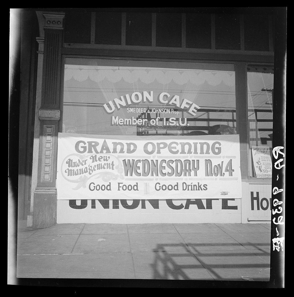 A black and white photograph captures a moment outside the Union Cafe, showcasing two large banners on its storefront. The top banner reads "Union Cafe" with additional text underneath indicating membership in I.S.U., which appears to stand for Industrial Section of United Order or another organization related to labor movement during that era.

Beneath this is an announcement sign reading "GRAND OPENING Wednesday Nov 4," underlining a significant event marking the establishment's inauguration. The advertised highlights include 'Good Food,' 'Good Drinks' and mention of 'Under New Management.' Another banner displays support for new management with logos from Smedler & Johnson Prop., hinting at their connection to I.S.U.

The setting is urban, as indicated by the sidewalk, building structure, and reflections in a window pane. The photograph has an air of historic significance, potentially dating back to mid-20th-century America based on its style and textual content which references unions commonly associated with labor movements during that time period.