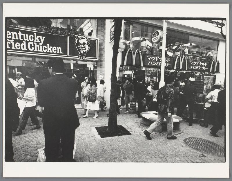 The image is a black and white photograph depicting an urban street scene with people walking around in front of fast food restaurants. In the foreground, there's a man dressed formally wearing glasses from behind, looking to his left as he walks. To his right stands another figure facing away from us; their attire suggests they may be part of security or staff. The middle ground is occupied by several individuals engaged in various activities: some walking towards and others away from the fast food outlets.

On the left side of the photo is a prominent Kentucky Fried Chicken (KFC) restaurant with its iconic branding displayed prominently above the entrance, featuring an illustration of Colonel Sanders. Next to it on the right stands another establishment bearing the golden arches of McDonald's, where people are queuing and ordering at what seems like a window service.

The architecture includes storefronts with large glass windows reflecting the street activity within them. A tree trunk is visible in the center foreground, providing some contrast against the urban setting. The sidewalk appears to be made up of cobblestones or bricks. On the right side near the curb, there's an open manhole cover on the pavement.

The overall scene captures a bustling atmosphere with people going about their day around these two fast-food giants in Japan during what seems like late 20th-c [...]
