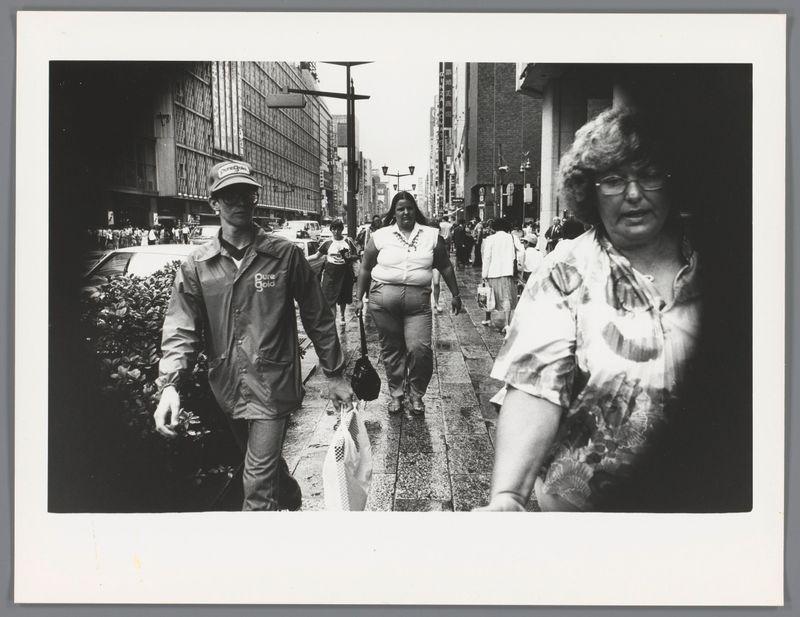 The image is a black and white photograph depicting an urban street scene with several pedestrians walking in different directions. In the foreground, there's a man on the left side wearing glasses, a cap labeled 'Nike', and what appears to be a windbreaker jacket; he seems focused on his destination or perhaps lost in thought as he walks down the sidewalk. The central figure is an overweight woman dressed casually with a handbag over her shoulder, walking confidently ahead of him.

Behind these two individuals are other pedestrians going about their business. Some wear casual clothing while others have more formal attire, indicating diversity among city dwellers captured at this moment in time. Buildings line both sides of the street, showcasing commercial establishments and urban infrastructure typical for a busy downtown area during daylight hours.

The photograph exudes an air of candid realism with its unposed subjects caught mid-movement within their daily routines, emphasizing life's hustle and bustle on city streets without any specific focal point or subject. The image is reminiscent of the work by Ed van der Elsken known for his street photography in Japan during a certain period.

The photo has an identifiable edge at each side suggesting it may be part of a larger collection or series, as indicated by the white borders framing this particular snapshot within w [...]