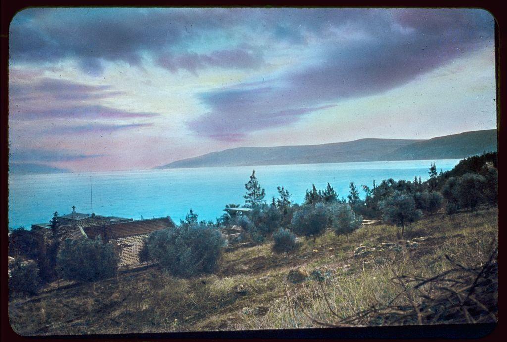 The image depicts a tranquil evening scene at the shores of Lake Galilee, also known as Kinneret in Hebrew. The perspective is from an elevated vantage point overlooking a picturesque landscape featuring lush greenery and trees with dark silhouettes against the lighter sky.

In the foreground lies a quaint stone structure perched on uneven terrain covered by dry grasses and shrubs. This building appears to be part of a larger facility, potentially associated with historical or religious significance given its isolated yet prominent location overlooking calm waters.

The central focus is Lake Galilee itself, displaying soothing hues ranging from deep turquoise near the shorelines to lighter shades where it meets the sky. The water reflects hints of pink and blue, suggesting either sunset colors or atmospheric reflections. A small boat or vessel can be seen in the distance on the lake's surface, adding a sense of scale.

Beyond the lake, rolling hills form a gentle backdrop under an expansive twilight sky painted with soft gradients from purple to yellow. Wispy clouds are scattered across this vast expanse above, contributing to the overall peaceful ambiance conveyed by the image.

The photograph carries historical connotations as it is identified within "Around the Sea of Galilee," documenting scenes in Israel between 1950 and 1977 through Matson Photo Service. The source  [...]