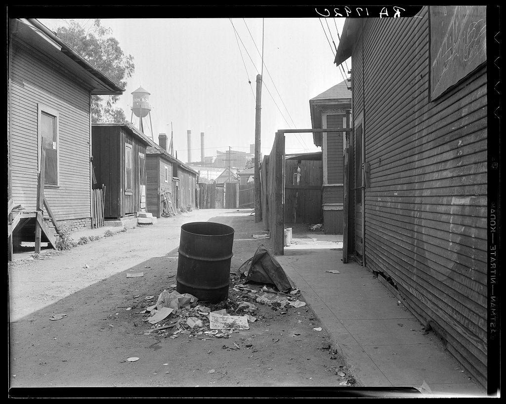The image depicts an urban alleyway with worn and weathered buildings on either side. There is a prominent barrel in the foreground, surrounded by scattered debris including pieces of wood and paper strewn across the ground. The alley appears narrow, with uneven pavement that has patches missing or displaced. Overhead power lines run parallel to one wall of the alley.

Various items are visible along the walls: what seems like crates or containers leaning against a building on the left side, an electrical box, and multiple pipes or cables running vertically up the sides of some buildings. There is also graffiti on one of the wooden surfaces, including words that read "HAIR" in large letters above smaller text.

In the background, several structures suggest this could be part of a working-class neighborhood with older architecture. Notably, there's a water tower labeled “CITY WATER” and what appears to be industrial smokestacks or chimneys further back, indicating proximity to an industrial area. The sky is overcast, contributing to the overall gritty ambiance of the scene.

The image carries historical significance as it was taken in Los Angeles' Mexican quarter near City Hall, highlighting aspects of urban decay within a densely populated community during that period.