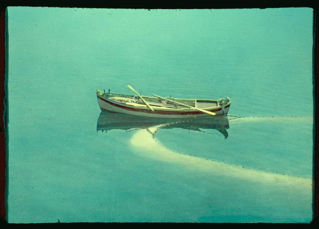 This image depicts a small boat floating on calm, turquoise waters. The vessel is painted white with red stripes and appears to be a simple motorboat rather than a traditional sailing craft or rowboat. There are no people visible in the photograph, giving it an abandoned feel as if the boat has been left there for some time.

A notable feature of this image is that its colors have faded over time; the overall hues appear washed out and lack vibrancy compared to what might be expected from a modern photo. The water around the boat displays a trail-like reflection known as "loop" or drag-net, which indicates recent movement within an otherwise still body of water.

The photograph carries historical context, with additional information provided in text that states it is taken between 1950 and 1977 at Bethsaida near the Sea of Galilee. This region has been a place for significant religious events and archaeological discoveries throughout history. The source "Matson Photo Service" suggests this image was captured by a professional photography company, which could be related to Matson Lines Inc., an American shipping line based in Honolulu.

Overall, the photograph conveys a sense of peacefulness with its serene waterscape and evokes curiosity about the boat's story within that historical context.