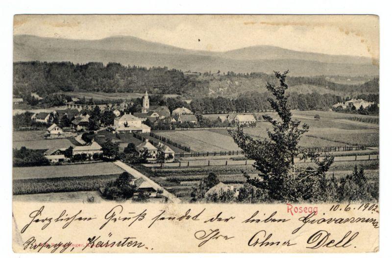 The image depicts a black and white photograph, seemingly from the early 20th century based on its style. It shows an expansive rural landscape with rolling hills in the background under a cloudy sky. In the foreground is handwritten text at the bottom of the photo which appears to be signatures or dedications along with some non-Latin characters suggesting it may have been taken by someone familiarly associated with Rosegg, Austria. The town within the photograph has several buildings including what looks like residential homes and possibly an institutional building with a prominent tower resembling that of a church or schoolhouse in its architectural style.

The landscape includes fields with trees scattered throughout, typical of rural farmland areas. There is also another handwritten inscription on top right which seems to be a date "Rosegg 10-6-1923", possibly indicating when the photo was taken or sent from this location.

Overall, it's an old postcard-like image showcasing both nature and human habitation in harmony with each other.
