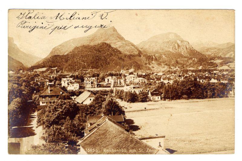 The image is a sepia-toned, vintage postcard depicting a panoramic view of the village Reichenhall in Bavaria. The photograph captures the settlement nestled amidst lush green hills and rugged mountains that stretch into the background under an overcast sky.

In the foreground, there's a cluster of buildings with varying architectural styles indicative of different periods or purposes - some appear residential while others could be commercial establishments like shops. A prominent church spire rises above other structures in the center, suggesting its importance to the community.

The middle ground is filled with more housing and possibly larger institutional buildings such as schools or administrative offices. The village seems organized yet spread out across a valley surrounded by greenery that indicates either farmland or forested areas suitable for agriculture.

In the background, majestic mountains rise steeply from the slopes of rolling hills, their rugged forms softened slightly by atmospheric perspective which makes them appear hazy with distance. This mountainous backdrop gives a sense of isolation and tranquility to this picturesque scene.

On top of the postcard is handwritten script that appears decorative, possibly indicating a dedication or ownership mark reading "Galia Heliaine T.V., Origine pur angure". Below in print, it states "1385 b. Reichenhall von St [...]