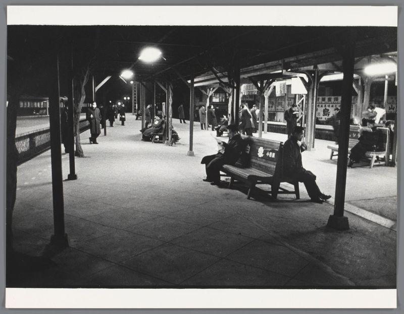 The image is a black and white photograph depicting an outdoor scene at night. It shows several individuals waiting on a platform, presumably in Japan given the caption reference. A prominent feature of this photo is its composition: multiple people are seated or standing under what appears to be a covered area with benches placed along it.

In one corner, two men sit facing each other; both wear dark coats and hats typical of mid-20th-century fashion, suggesting a historical context for the image. Another man sits on a bench in profile view, reading something that resembles newspapers or magazines. The platform extends into the background with more individuals visible, some walking along the edge.

Street lamps illuminate the scene, casting shadows and highlighting textures such as wooden beams supporting the structure overhead. A train is partially visible under this canopy at one end of the photo, which reinforces the setting's transportation context. There are signs hanging from above; however, they're not legible in the image provided. The overall mood conveyed by the photograph suggests a momentary pause or daily routine among people waiting for their transport during nighttime hours.

The caption mentions "Reizigers wachten op een perron" which translates to 'Travelers wait at an platform,' and credits Ed van der Elsken as the photographer, with years 1959-60 indic [...]