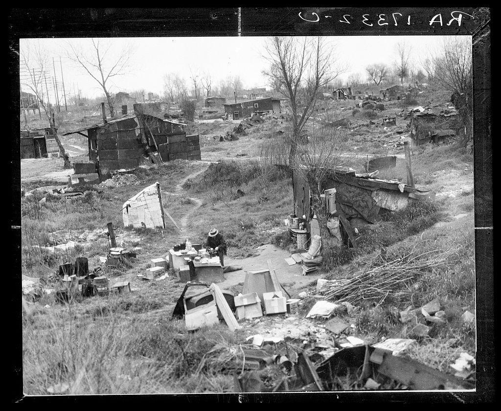 The image depicts a desolate, rundown area with makeshift structures made from various materials such as wood and metal. There's an overall sense of abandonment or neglect in the environment. The black-and-white photograph captures the scene on what appears to be overcast day given the absence of strong shadows.
In this setting, there is one individual sitting amongst these ruins. This person seems to be seated outside a small shanty-like structure and has their back partially turned towards the camera, making it difficult to discern any facial features or expressions from that angle alone. The surrounding area includes scattered debris such as broken pieces of furniture, discarded household items like buckets and cans, piles of branches, and other assorted refuse.
The overall atmosphere conveyed by this image is one of hardship and decay, suggesting a lack of proper living conditions for the inhabitants in what appears to be an impoverished or marginalized community. The photograph seems reminiscent of historical social issues regarding housing and displacement faced by certain demographics during that era.