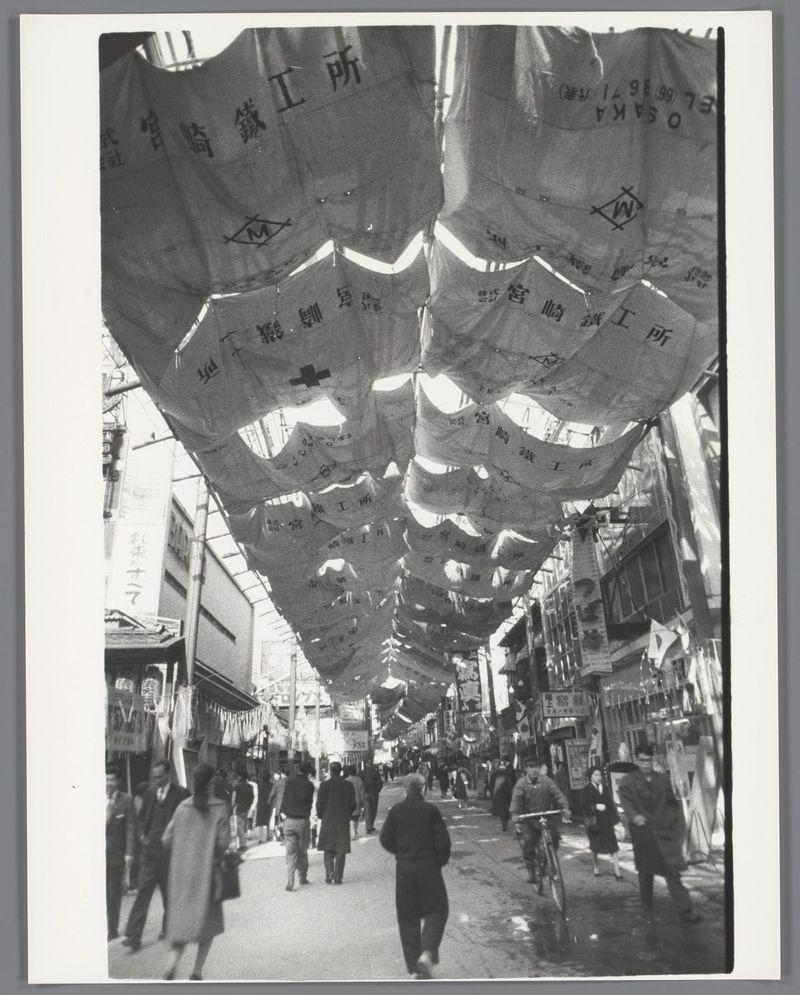 The image displays a bustling urban street filled with pedestrians, captured in monochrome. The scene is dominated by large fabric banners suspended overhead, creating an arching canopy over the walkway. These banners bear Japanese characters and symbols such as medical crosses, suggesting they may be associated with healthcare or community organizations. People of various ages can be seen walking beneath these banners; some are dressed formally while others wear more casual attire. The street is lined with what appears to be shops or market stalls on both sides, indicating a commercial area likely in an Asian city. Light filters through the gaps between the hanging fabrics, illuminating parts of the scene and casting shadows elsewhere.