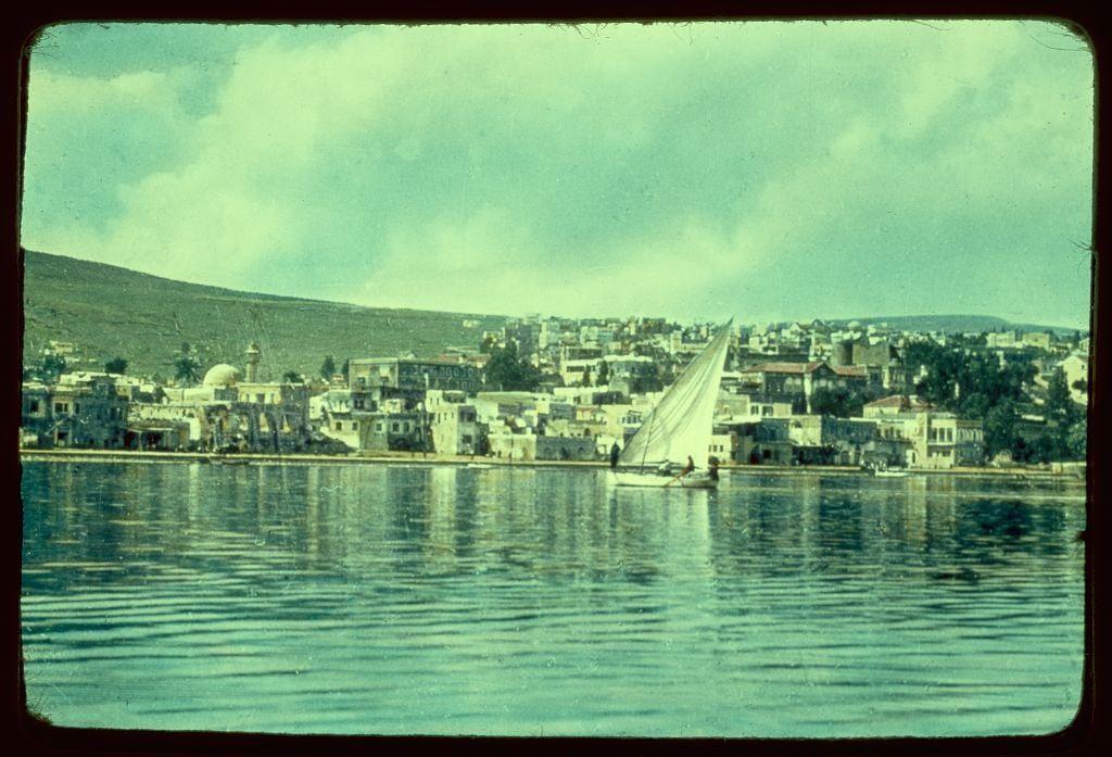 The image depicts a picturesque coastal town with buildings lining the shore, extending into the distance. A sailboat is floating on calm waters in the foreground, its white sails billowing slightly as it appears to be moored or moving slowly. The sky above is mostly clear with some scattered clouds, and there's a gentle slope leading up from the water towards residential areas that fade into greenery further inland. This setting suggests a peaceful Mediterranean town, perhaps near a significant body of water like the Sea of Galilee mentioned in its context.

The photograph has an old-fashioned aesthetic, likely taken during or after World War II given the dated quality and color saturation typical of mid-20th-century photography. The image is credited to Matson Photo Service, indicating it's part of their collection of photographs documenting various locations over a period of years.