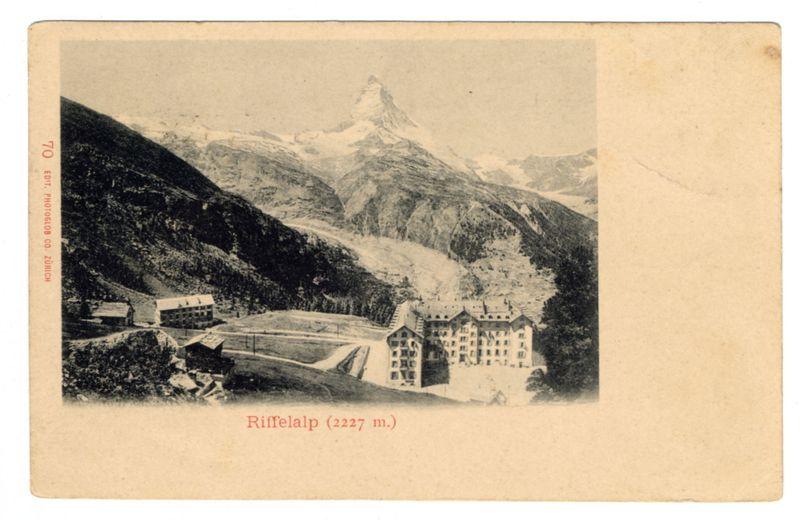 The image is a black and white photograph with an aged appearance, suggesting it may be quite old. It features the Riffelalp landmark at 2227 meters above sea level, as indicated by text in red on the bottom of the photo. The landscape prominently displays mountains covered with snow or ice, indicating cold temperatures typical for high-altitude locations.

In the foreground and middle ground, there is a large building complex which appears to be an alpine hotel or resort named Riffelalp. This structure has multiple stories, numerous windows, and what seems like several wings extending outward. The architecture of the buildings suggests they were constructed with functionality in mind for accommodating visitors.

Surrounding this main edifice are smaller structures that could serve various purposes such as offices, storage facilities or additional guest accommodations. There's a noticeable presence of infrastructure on the slopes leading up to Riffelalp; these might be roads, paths, or perhaps even cable car tracks used by tourists and workers alike.

The lower left corner has text in red which says "70" with what seems like an edit notation below it followed by some non-Latin script that could indicate a language such as Russian. This suggests the photo may have been part of a series collected for publication or archiving purposes.

Overall, this image captures not just  [...]