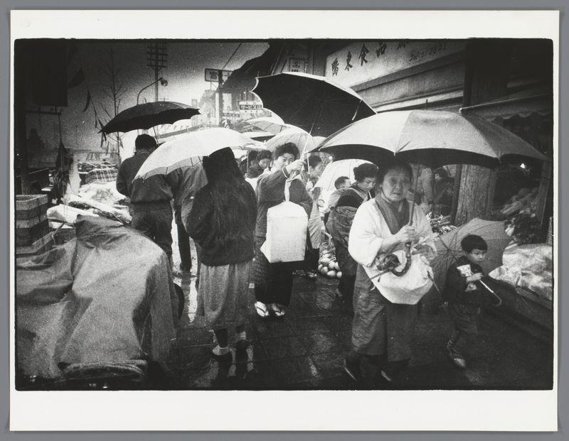 The image captures a bustling street market scene on what appears to be a rainy day. Multiple people are navigating the area, with several holding umbrellas over their heads, suggesting precipitation. The crowd includes men and women of varying ages, some wearing traditional attire that suggests cultural significance or possibly participation in an event like a festival. There's visible movement as individuals walk through narrow market aisles filled with stalls displaying goods for sale. The photograph is monochromatic, which adds to the historical atmosphere, likely placing it from early photography days when color film was not prevalent. Notable features include street signage written vertically on buildings in East Asian characters, indicating a setting in Japan or another Japanese-speaking region. There's also a sense of everyday life and commerce captured here, with people going about their activities despite the weather conditions. The image conveys a dynamic yet candid snapshot of daily urban life in an Eastern country during what seems to be the mid-20th century based on fashion styles and infrastructure.