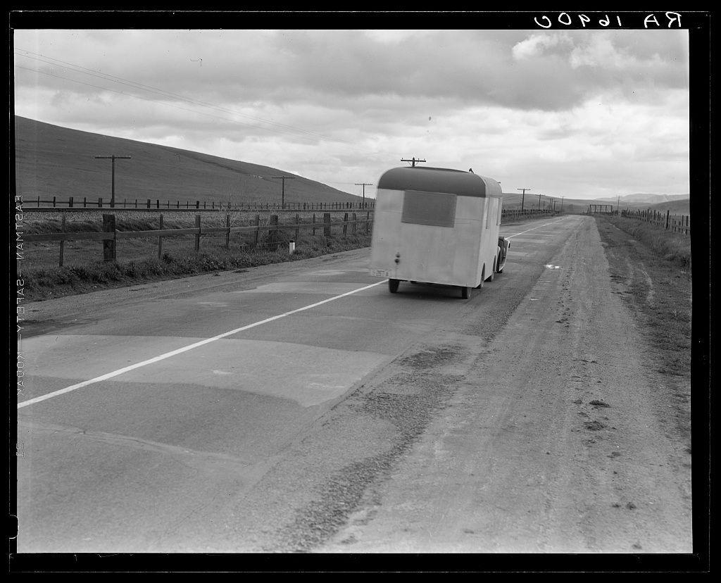 The image is a black and white photograph depicting an old-fashioned trailer being towed by another vehicle on what appears to be an expansive rural road. The scenery suggests it could be in the western United States, with open fields, fences running parallel alongside the highway, and rolling hills visible in the distance under a cloudy sky.

In terms of alt text for visually impaired users:
- A black and white photograph showing a vintage trailer being towed on U.S. 101 near King City.
- The image captures an older couple likely living out of their homemade trailer as they journey from Wisconsin to California during tough economic times, suggesting the challenges faced by people in mobile housing during that period.

Additional context provided through notes indicates:
This picture is linked with a personal story about two middle-aged individuals who left on the road due to unemployment (referred to humorously as "Old Man Depression"). They have been traveling for around two years and share their experiences of encountering various situations involving people living in trailers, particularly when they arrived at Florida.