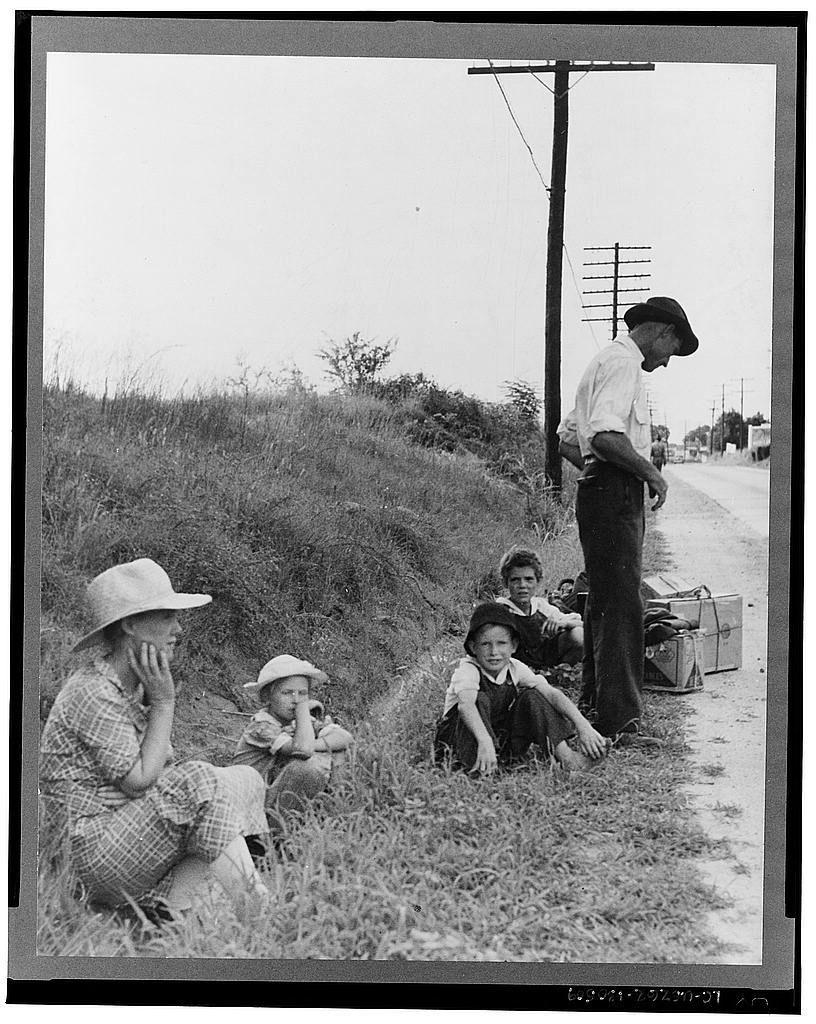 The image depicts a family of five individuals situated by the side of a rural road. The setting is outdoor, with overcast skies and vegetation in the background that appears unkempt or wild. A telephone pole stands prominently on the right-hand side.

In terms of age distribution within the group:
1. One adult male, presumably the father.
2. Two adolescent boys.
3. Three younger children (two girls), one seated close to an older child who seems to be in a protective stance and another kneeling beside them with hands near their mouths possibly indicating distress or contemplation.

Their attire suggests modest living conditions; it is simple yet functional for outdoor work, reflecting the rural context of Macon Georgia where such family would reside. The clothing varies from plaid dresses, shorts, caps, to shirts and trousers, all appearing well-worn but practical.
The overall mood captured seems pensive or weary as they await their next step in life's journey marked by uncertainty indicated through body language like hand on face, leaning posture among adults while the children present a range of expressions from contemplation to distress.