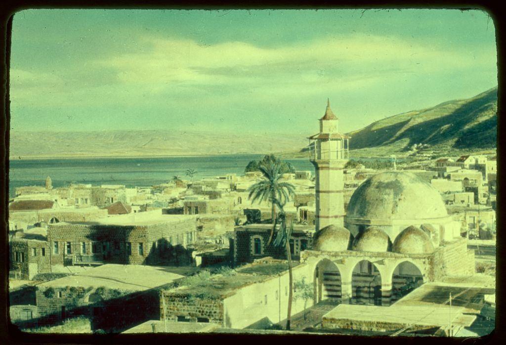 The image depicts a panoramic view of Tiberias, Israel, showcasing the ancient architecture and historical buildings that characterize this region. Prominent in the center is an Islamic mosque with its distinctive dome and minaret standing out against the urban landscape. Surrounding structures appear to be built from stone or stucco, indicative of traditional Middle Eastern construction techniques.

In the foreground, a palm tree stands tall amidst the rooftops, adding a touch of greenery to the otherwise earth-toned scene. The sky is partly cloudy with hues ranging from light blue to soft yellowish-green, suggesting either early morning or late afternoon lighting conditions. In the background, rolling hills are visible along the shoreline of what appears to be the Sea of Galilee, providing a serene and picturesque backdrop.

The image has an aged look, likely due to its vintage origins as part of Eric and Edith Matson's photo collection from around 1950-77. The photograph captures not just the physical environment but also offers insight into the cultural heritage of Tiberias during that period.