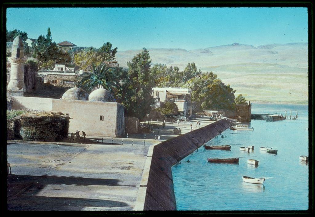 The image depicts a picturesque waterfront scene, likely captured during the mid-20th century. It features serene blue waters with multiple small boats scattered across them, suggesting an active harbor or fishing area. In the foreground is a wide walkway along what appears to be a defensive wall, leading towards a fortified building complex that includes domed structures and other architectural elements typical of Middle Eastern design. This setting likely represents Tiberias on the Sea of Galilee.

The background reveals rolling hills with sparse vegetation under a clear sky. There are people visible in various parts of the scene, indicating daily life activities near this waterfront area. The overall atmosphere conveyed is one of tranquility and historical significance, captured by Matson Photo Service as part of their collection documenting locations around the Sea of Galilee between 1950 and 1977.