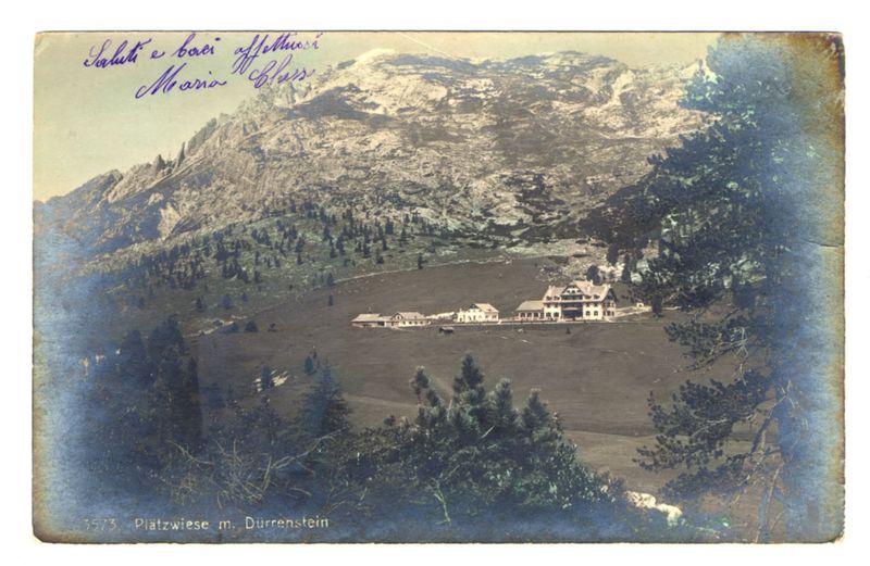 The image displays a vintage postcard featuring an expansive mountainous landscape with snow-capped peaks in the background. In the foreground, there is a lush green valley dotted with trees and coniferous forests. Nestled within this picturesque setting are several grand buildings reminiscent of alpine chalets or perhaps early 20th-century resorts. The architecture suggests European influences, likely Germanic due to the context provided by "Plätzwiese m. Dürenstein," which could be translated as a pasture with Dürrenstein. Atop these structures, prominent features include sloping roofs and large windows, indicative of designs suited for mountainous climates.

The overall coloration is sepia-toned, typical of aged photographic prints or postcards from the early 20th century. The edges show signs of wear, including fading and discoloration, hinting at its considerable age. Prominently featured in the top left corner are handwritten inscriptions: "Salute e buon viaggi" translates to "Greetings and safe travels," followed by a signature or name that appears difficult to decipher without context.

In the bottom right corner is an inscription providing location details with "3573 - Platzwiese m. Dürenstein." This likely denotes the card's catalog number within a collection, while also labeling it as being taken from Dürrenstein. The combination of these elements paints a vivi [...]