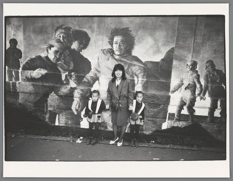 The image depicts a black and white photograph of three individuals standing in front of an outdoor mural. The mural is quite large, taking up the entire background wall behind them, with multiple figures depicted within it, appearing to be engaged in various activities or expressions.

In the foreground, there's one adult woman and two young girls dressed similarly, likely indicating a family connection such as mother and daughters. All three individuals are wearing jackets and skirts, suggesting cool weather attire typical of many East Asian countries during that time period. The girl on the left holds hands with her sister, both looking directly at the camera.

The mural itself seems to portray different scenes or characters; some appear older and more ruggedly dressed in coats and hats indicative of a colder climate. Others are shown seemingly in distress or hardship. There's also an element of motion suggested by one figure holding onto another as if being pulled away from something, adding drama or tension.

The setting appears urban with the pavement beneath their feet visible along with some litter suggesting public space like a street or pedestrian area. The photograph is framed within what looks to be a Polaroid-like border which adds an informal and personal touch to it. This image evokes themes of daily life juxtaposed against historical narratives represented [...]