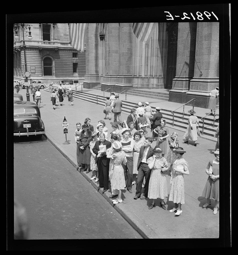 A black and white photograph showing people on the sidewalk in front of a large building, likely a church given its architectural style. There are several individuals standing or walking along the curb, with some looking at what appears to be maps or pamphlets. A woman wearing glasses is holding papers up for others around her to see. Several cars can be seen parked and driving on the street next to them. The image has text overlay in the top right corner that reads "3-2189" along with additional numbers, possibly indicating a date code.