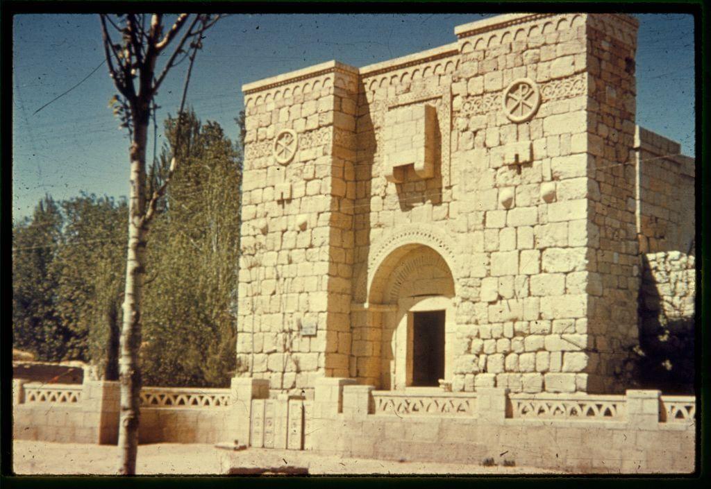 The image depicts a historic stone structure, likely part of an ancient architectural complex. The building features two prominent towers with circular emblems or crests above them. Between the towers is a large arched entrance flanked by smaller arches and decorative stonework.

In front of the building stands a leafless tree on the left side and a clear blue sky in the background, indicating that it's likely taken during a sunny day. The setting appears to be outdoors with some greenery visible around the structure.

The image has a vintage look, suggesting it may have been captured using film photography or processed for an aged effect. There are also signs of wear or damage on the photo itself, such as scratches and discoloration along the edges.

Additional context provided indicates that this location is significant in historical research, specifically mentioning Damascus, Palmyra, and Baalbek sites related to archaeological studies during a restoration period between 1960-1977. The photograph was taken by Matson Photo Service as part of their collection or archival materials.