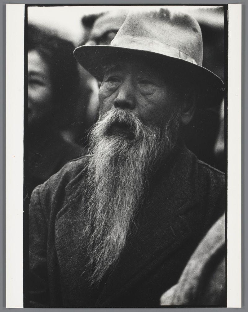 The image is a black and white photograph showing an elderly man with a long, full beard extending down to his chest. He wears a wide-brimmed hat that casts shadows over the top of his head. The man appears contemplative or perhaps in awe, as suggested by his slightly open mouth and upward gaze directed off-camera's left side. His eyes are not visible due to the angle at which he is looking away from the lens.

He has deep-set wrinkles on his forehead and under his eyes, indicating advanced age. He wears a dark, textured garment that could be a coat or jacket over what seems like another layer of clothing underneath. The background is indistinct but appears crowded with other figures partially visible behind him, suggesting he may be in a public setting.

The style of the photograph has a documentary feel to it, capturing an authentic moment rather than a posed portrait. There are no discernible colors due to the black and white medium; however, there's contrast between light and shadow that defines textures like his beard or hat material effectively.