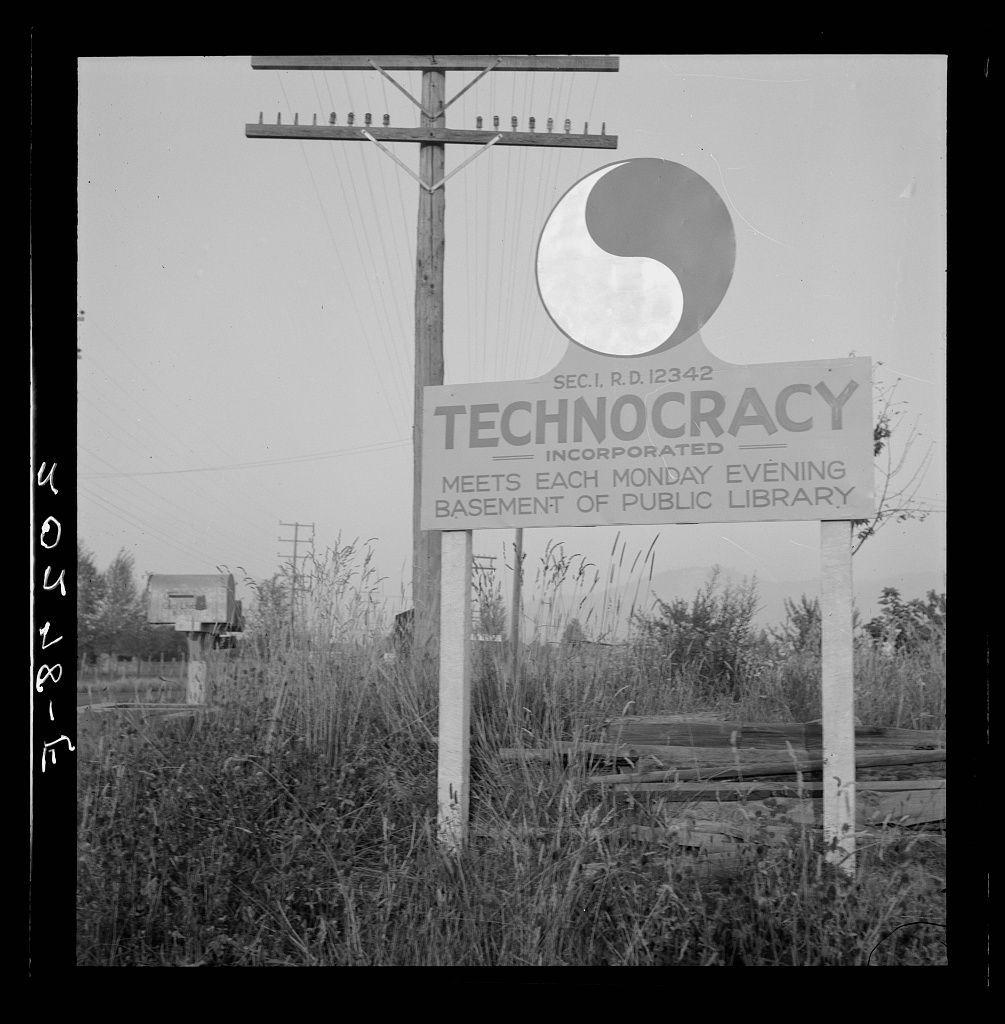 The image is a black and white photograph depicting an outdoor scene. The focal point of the photo is a large sign that reads "TECHNOCRACY INCORPORATED" with additional text stating it meets each Monday evening at the basement level of the Public Library. Above this main title, there's a symbol resembling a Yin-Yang, indicating some connection to balance or duality in ideology.

The background shows an overcast sky and what appears to be tall grasses swaying gently in the breeze. There are utility poles with attached lines running parallel to each other across the top part of the image. The environment suggests it could be on the outskirts of a town, as there's no visible urban infrastructure beyond these natural elements.

A wooden mailbox is also present beside the signpost, further emphasizing this rural or semi-rural setting. There are some fallen and weathered boards at the base of one pole which seem to have been repurposed for something else. The overall scene conveys a sense of abandonment yet purposeful symbolism with its prominent sign.

The photo seems historical in nature given its black-and-white format, suggesting it could be from an earlier time period when such gatherings or groups were active and perhaps more common.