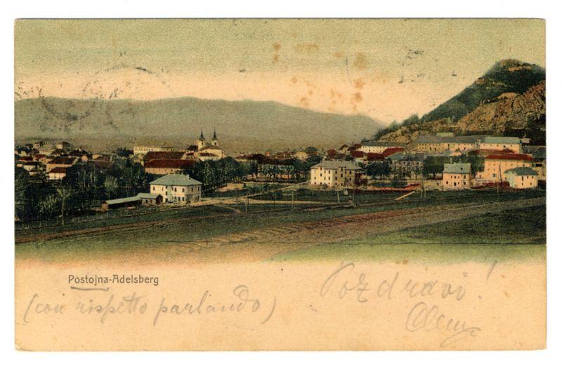 The image depicts a vintage postcard showing an aerial view of a town nestled amidst rolling hills. The prominent feature in the landscape is a large mountain or hill to the right, suggesting that this location could be situated near elevated terrain. Below it, there's a collection of buildings with varying architecture styles and roof colors ranging from red tiles to slates. A few notable structures include what appears to be an institutional building at the forefront due to its size and design, possibly a school or church based on its architectural features.

The town is set in a valley surrounded by low-lying hills that fade into the distance under a cloudy sky with soft hues of pale blue and white. The colors are muted but convey a sense of tranquility typical for historical postcards from this era.

On the left side, there's handwritten text indicating an address or destination: "Postaja-Adelsberg" followed by what seems like a misspelled phrase "-inadispolto parlavio," and another fragment that reads "-cudrau." On the right-hand side, in cursive handwriting, it says "Coedruai Cuyy."

The overall condition of the postcard suggests age with some spots and stains visible on its surface. The text at the top left corner is printed: “Postoja-Adelsberg (coi hispetto parlavio) Coedrui!” indicating a potential location or destination, which seems to be in an Italian-speaking [...]