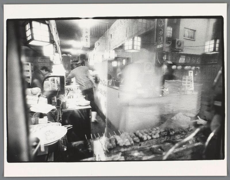 The image captures a bustling, monochromatic scene within what appears to be an Asian street food market or restaurant. There is a sense of motion conveyed through the blurred figures and objects which suggest rapid movement and activity. In focus are various elements: stacks of plates awaiting use at a serving counter, skewers likely containing grilled meats over a grill, and patrons engaged in dining activities—some seated while others stand. The background features signage with Japanese characters suggesting this setting is indeed in Japan or caters to that demographic. Overhead lighting fixtures emit bright light onto the scene below. Notably absent are vibrant colors; instead, shades of gray dominate due to the black-and-white film used by photographer Ed van der Elsken during his time capturing urban life and culture across Asia from 1959-1990.