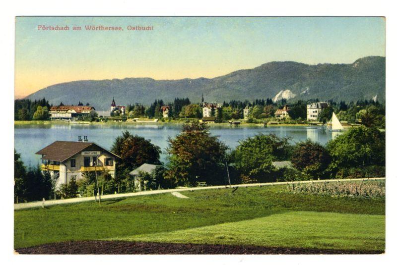 The image is a vintage postcard depicting the picturesque lakeside scenery of Portschach am Wörthersee in Ostbucht, Austria. The scene showcases calm waters reflecting shades of blue and green under a light sky with distant mountains providing a backdrop. A variety of buildings are scattered around the shoreline; some resemble traditional alpine houses with sloping roofs and others appear more modern or institutional. One such building is prominently located on the lakeside, possibly an inn or lodge, distinguished by its prominent red roof.

Trees line the shore, adding greenery to the landscape, while well-maintained lawns occupy the foreground of the image. A small sailboat with a white hull and mast is visible in one corner of the lake, suggesting recreational activities are enjoyed here. The composition includes text at the top left that reads "Portschach am Wörthersee, Ostbucht," likely indicating both the location and perhaps its Italian name.

The overall ambiance conveyed by this image suggests a serene vacation spot with an emphasis on natural beauty and relaxation.