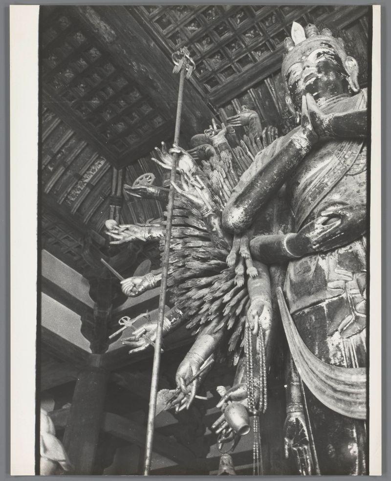 The image is a black and white photograph capturing an intricately detailed statue, likely of religious or cultural significance. The central figure appears to be Kannon, also known as the Bodhisattva of Compassion in Buddhism, depicted with multiple arms extending outward holding various symbolic items such as flowers, beads, and a sword. Each arm is meticulously sculpted, showcasing fine craftsmanship indicative of traditional Japanese artistry.

The statue's face expresses serenity amidst complexity, suggesting a revered spiritual presence within a temple or sacred space. The background reveals architectural elements typical of East Asian temples, with wooden beams featuring ornate carvings that complement the statue’s intricate details.

There is also a person standing beside the statue, though not in focus and partially obscured by shadows, possibly indicating their reverence towards this deity during worship or observation within the temple setting.