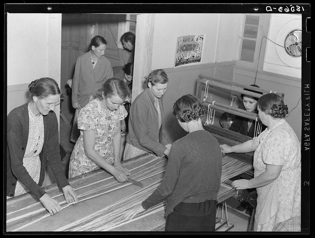 The image depicts a group of women engaged in rug-making activities within what appears to be an industrial setting. The focus is on five female workers who are seen handling large pieces of fabric that seem to form part of the rug or carpet being produced. They wear various garments, including dresses and cardigans, suggesting a casual work environment. One woman stands out as she interacts with a machine likely used for weaving or assembling parts of the rug.

The background reveals more individuals involved in different tasks within this workshop-like space. A sign on one wall reads "HAVE A HOBBY," indicating an emphasis on recreational activities alongside their labor. The image has a historical feel, possibly from mid-20th century America, judging by clothing styles and overall setting. No explicit logos or identifying marks are visible to ascertain the exact location of this scene.

The photograph captures not only the women's industrious task but also reflects broader societal themes such as gender roles in industry during that period and communal activities within a migrant camp environment, hinting at collective efforts for livelihood and education among the community.
