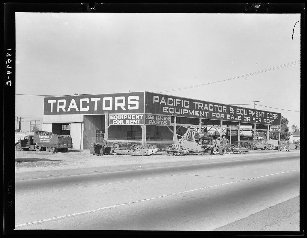 This black and white photograph depicts a bustling industrial setting from an era in the past. The focal point is a large, prominent sign that reads "TRACTORS" above another larger banner stating "PACIFIC TRACTOR & EQUIPMENT CORP." Additional text on this banner indicates offerings of equipment for sale or rent, including tractors and parts, as well as mentions of used equipment availability.

The business appears to be situated alongside a road with a clear demarcation line in the middle. Vehicles are visible at various distances from the camera's viewpoint; closer to the building is an older model truck marked "SUGAR OIL CO." parked directly against it. Other cars and trucks can be seen scattered across what seems like a lot filled with construction machinery, including cranes and other heavy equipment.

The sky above appears clear but devoid of any distinctive features that might indicate weather conditions or time of day beyond the general daylight setting suggested by the image's lighting. The overall impression is one of an active commercial enterprise engaged in selling farm-related machinery during what could be a significant period for such businesses, possibly related to agricultural activities in California as indicated by the reference text.

The photograph conveys not just an industrial scene but also hints at economic activity and perhaps reflects on histori [...]
