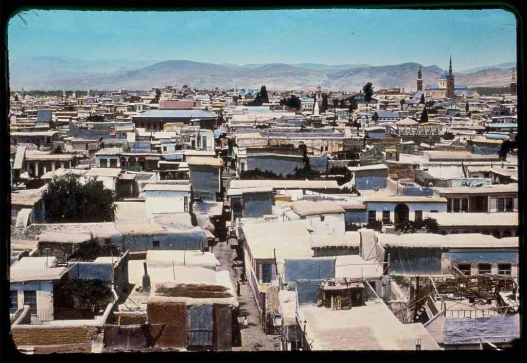 The image depicts an aerial view of a densely packed urban area, showcasing numerous buildings with varying architectural styles and roof designs. The structures appear closely built together in rows, creating narrow alleys between them.

Dominating the skyline is a large mosque with a prominent minaret reaching into the clear blue sky, indicating its religious significance within the community. In the background are mountains or hills under a bright sunlit day, adding to the picturesque quality of the scene. The overall impression suggests that this image was captured in Damascus during mid-20th century.

The photograph is framed with what seems like an old film frame on the edges, giving it a vintage appearance and hinting at its historical context as part of Eric and Edith Matson's collection from 1950 to 1977. The photo likely serves as a visual record of daily life in Damascus during that period, reflecting architectural styles, urban planning, and perhaps socio-economic conditions prevalent then.

Further information about this image can be found through references such as "Damascus," "Palmyra," and "Baalbek" within the provided sources or by searching for Matson Photo Service archives.