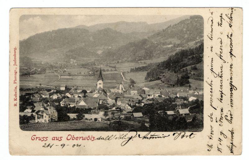 The image displays an antique postcard from Judenburg, Austria. The scene captured on the card shows a picturesque village with numerous buildings nestled among hills and surrounded by open fields. Prominent features include a central church spire flanked by other smaller structures that suggest residential housing.

At the top of the card, in red script text against a white background, it reads "Gruss aus Oberwölz," which translates to "Greetings from Oberwölz." Below this greeting is additional handwriting and signatures. The bottom left corner bears the name R. Kuballa with the initials P., Judenburg.

The overall aesthetic of the postcard suggests an early 20th-century origin, capturing a serene rural landscape typical of Austrian countryside during that period.