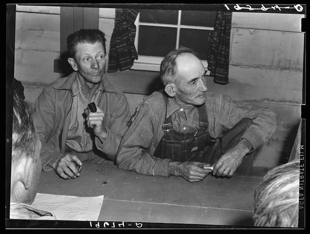 The image is a black and white photograph depicting two men seated at what appears to be an informal meeting or council, with the setting likely in California during the Great Depression as part of Farm Security Administration (FSA) documentation on migratory agricultural workers. The man on the left holds a small object which could possibly be a pipe, indicative of contemplation or concern. His counterpart is engaged in examining what seems to be paper documents placed before him.

The setting includes a rough-hewn wooden structure with visible planks and windows dressed with curtains that suggest modest living quarters typical for agricultural workers during this period. The photograph captures the candid interaction between these individuals without any formal attire, emphasizing their rural and working-class status. There is also an indication of ongoing discussion or deliberation as suggested by one man's pointed finger.

The image serves to provide a historical glimpse into migrant labor conditions in agriculture through a FSA documentation photo, which aimed at highlighting social issues faced during the Great Depression era across America.