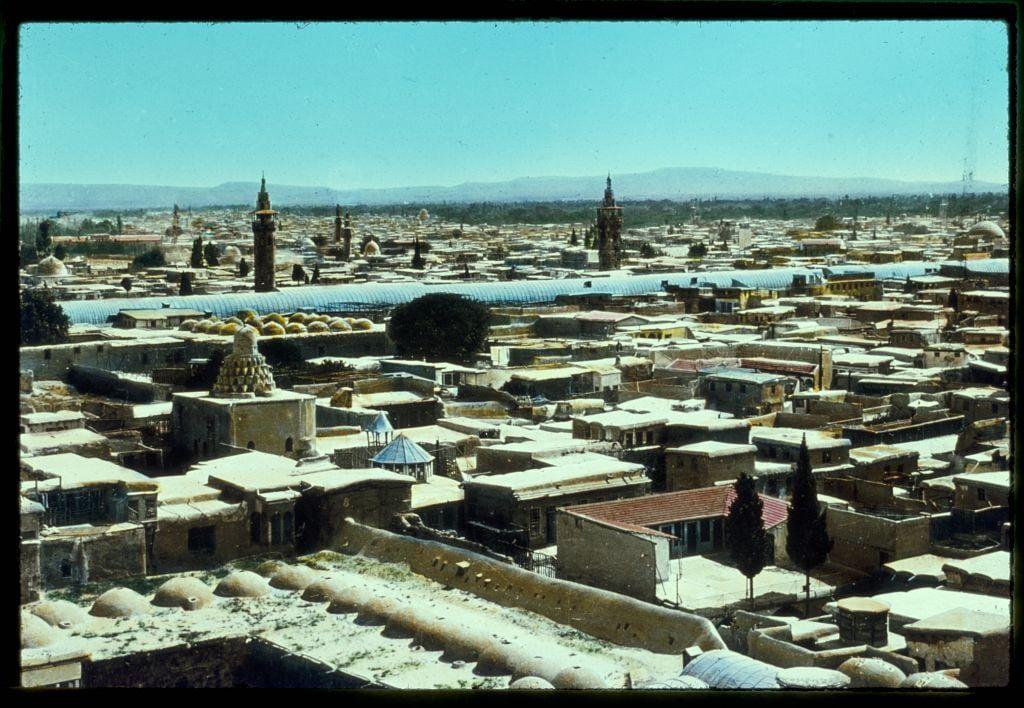This image displays an expansive aerial view of a historic cityscape, likely taken from a high vantage point. The scene is dominated by numerous buildings with flat roofs and various architectural styles suggesting historical significance. Some structures feature arched doorways and windows indicative of traditional design.

Prominent in the background are several minarets and domed towers that stand out against the skyline, hinting at Islamic architecture which might be associated with mosques or religious sites. The color palette is warm, with earthy tones on the buildings contrasting sharply against a clear blue sky.

The perspective from above allows for an overview of the urban layout, revealing streets lined with smaller structures and open spaces where vegetation can be glimpsed in certain areas. In the distance, mountains provide a natural backdrop to the city, emphasizing its expanse within the surrounding landscape.