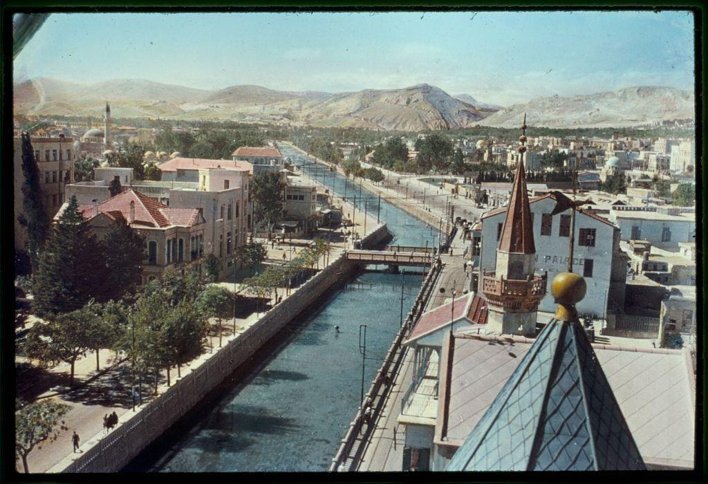 The image depicts a picturesque view of Damascus, showcasing the River Abana and surrounding architecture. It captures an aerial perspective that highlights colorful buildings with red rooftops along the riverbanks, various structures like mosques or churches with distinct spires, including one with golden dome and another resembling traditional Islamic architecture in its design.

In the background, mountains are visible under a clear sky, adding to the scenic beauty of this Middle Eastern city. The River Abana meanders through the urban landscape, bordered by trees and walkways where people can be seen strolling or engaging in daily activities.

The photograph is credited as part of an archive from Matson Photo Service, which suggests it could date between 1950 and 1977 based on provided information. The inclusion of such a historical image likely indicates its significance to the region's cultural heritage during that time period.