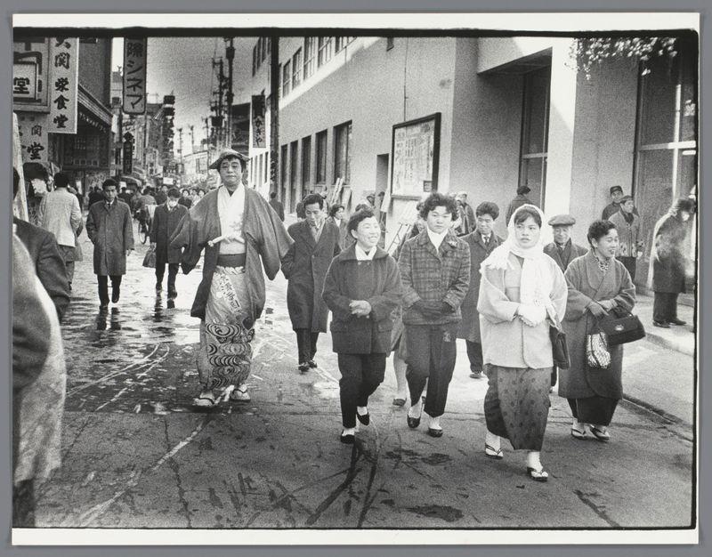 The image captures a bustling street scene in Japan, likely taken during the late 1950s to early 1960s based on its monochromatic style. The focus is on a group of people walking down the pavement; their attire suggests it may be autumn or winter as they are wearing coats and hats.

In the foreground, there's an individual dressed in traditional Japanese clothing that stands out due to its distinct patterns and flowing robes. This person appears to be leading the group. Behind this figure walk individuals who seem engaged with each other, indicative of a casual stroll rather than hurried movement. Some wear more modern Western-style clothes such as coats and trousers.

The street is lined with buildings bearing Japanese characters on their signage, suggesting commercial establishments like restaurants or shops. The urban environment indicates a city setting with the pavement wet from rain, implying recent precipitation. People are captured in mid-stride; some look towards the camera while others engage with passersby out of frame. There's no clear indication of any specific event other than it being an everyday scene.

This photograph is attributed to Ed van der Elsken and belongs to a series documenting Japanese life during that period, reflecting his interest in capturing candid moments within urban settings. His work often juxtaposes traditional aspects with modern ele [...]