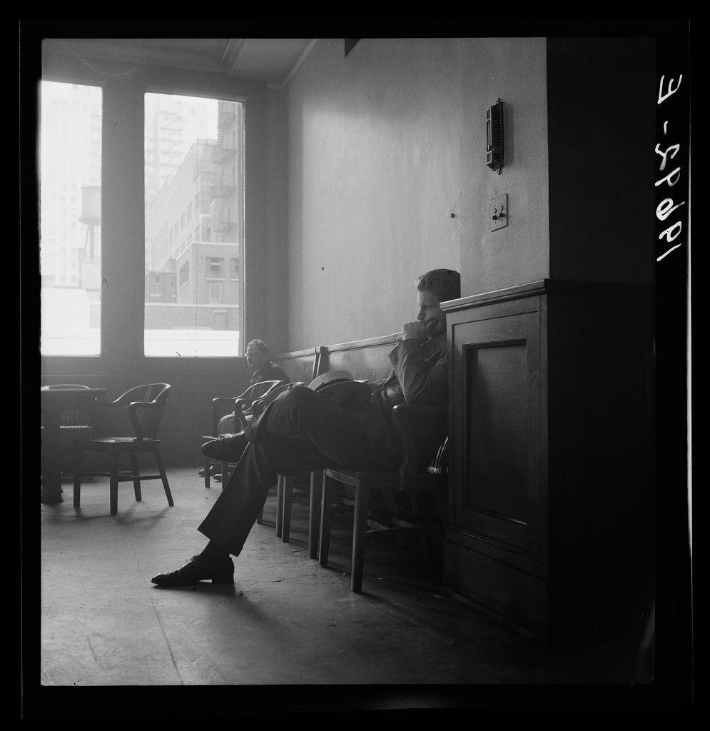 The image shows a black and white photograph depicting two individuals seated in what appears to be the committee room during a session of Chicago Board of Aldermen. The setting has large windows that allow daylight into the space, and an air conditioning unit is mounted on one wall above their heads.

One individual sits against a wooden cabinet or paneling with his back partially turned towards us; he rests his chin in his hand while looking out through the window. His legs are crossed at the ankles, wearing dark pants and shoes that suggest formal attire.

In contrast to him, another person is seated facing forward on one of the chairs, seemingly engaged with something off-camera or possibly waiting for an event. The room has a somewhat sparse furniture arrangement including wooden benches, empty chairs in front, and additional seating behind where there appears to be more individuals present but not visible due to angles.

The lighting casts shadows within the space, emphasizing the texture on surfaces like the flooring, which is composed of tiles or concrete slabs. Overall, this image captures a moment of waiting or contemplation during an official gathering in Chicago's municipal governance setting.
