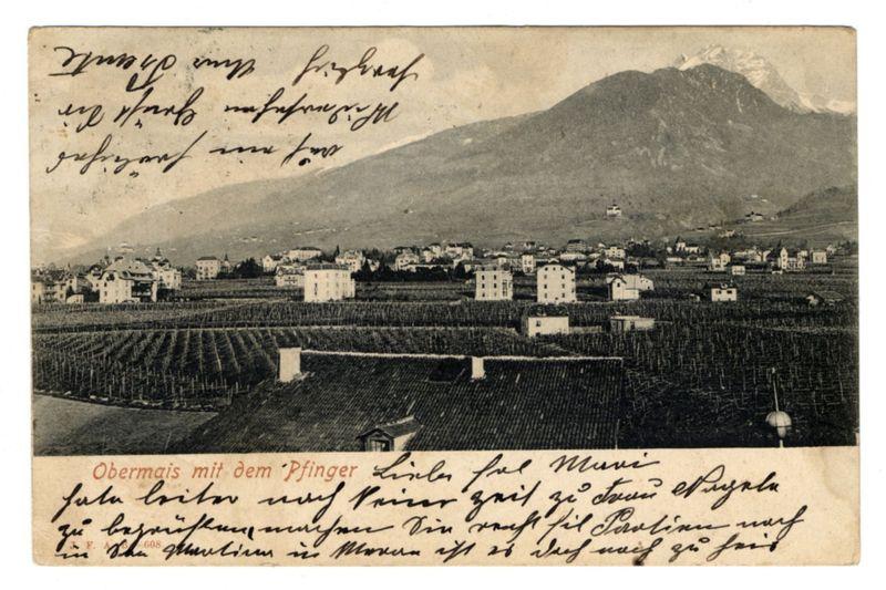 The image is a vintage postcard depicting an old European village nestled in the mountains. The landscape shows terraced vineyards stretching out across rolling hills, indicative of wine production common to this region's climate and geography.

In the foreground, there are rows upon rows of grapevines meticulously tended, suggesting meticulous agricultural practices typical for regions like Austria or northern Italy. In contrast, built-up areas feature a mix of buildings: residential houses with pitched roofs on one side and larger structures that could be wineries or storage facilities in the vineyards.

The mountainous backdrop is marked by snow-capped peaks under an overcast sky, providing a stark natural beauty juxtaposed against human habitation. The overall palette of the image is monochrome due to its vintage nature, with varying shades from white and grey for clouds and mountains to darker tones representing buildings and shadows on terraced fields.

The handwritten text in German overlaid at the bottom left corner suggests personal or historical annotations made by a postcard sender, perhaps commenting on different vineyards they visited. The handwriting is somewhat cursive and ornate, indicative of its age.

In terms of geographical context, Obermais could refer to various places worldwide but within European regions known for wine production like Austria's Bur [...]
