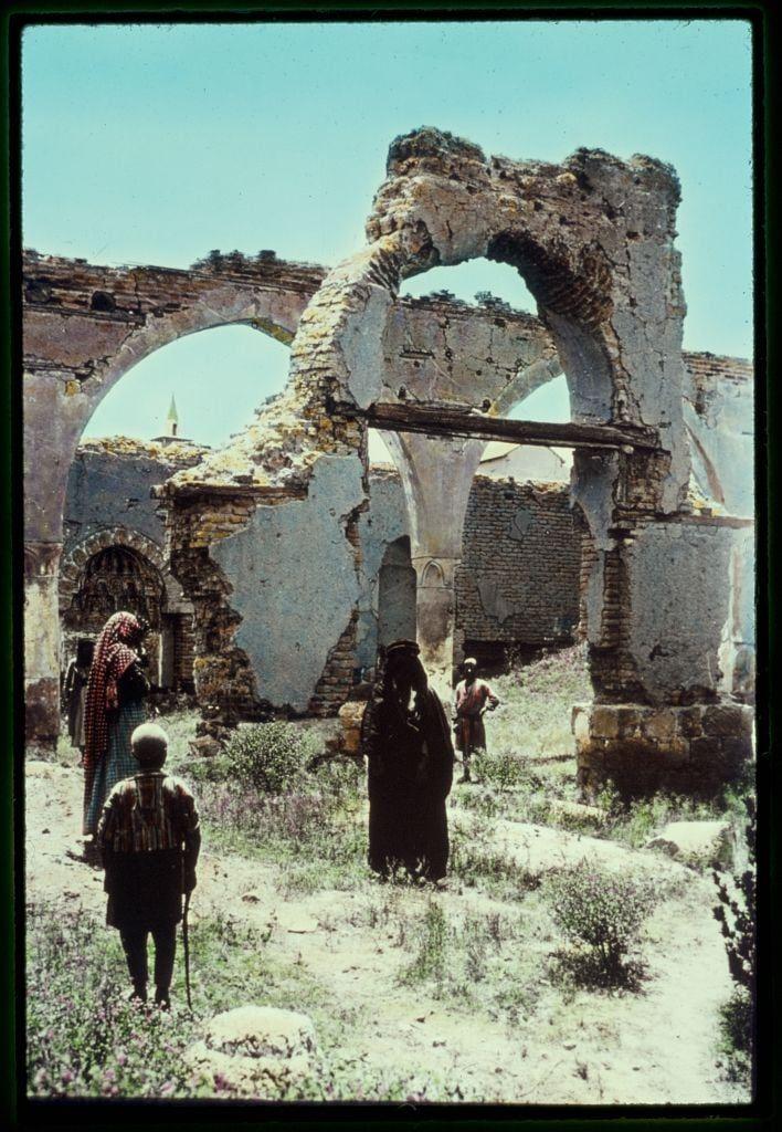 The image depicts a group of people walking through the ruins of an ancient structure. The setting appears to be arid and desert-like, with dry grasses and sparse vegetation around. There are three individuals visible: two women dressed in traditional attire - one wearing a red headscarf over her shoulders while holding what seems like bags or belongings, and another draped in black robes; the third person is an older man walking ahead of them with a cane for support.
The ruins themselves consist of large stone arches and walls that show significant decay and damage. The structure has multiple rounded openings at various heights, suggesting it could be part of a larger complex such as a temple or public building from historical times. In the background, there is another faintly visible dome-like shape with a green top which might indicate some other architectural element in the vicinity.
The overall scene conveys a sense of desolation and abandonment, capturing a moment where individuals are navigating through remnants of an old civilization's architecture amidst their daily lives or perhaps exploring the historical site.
