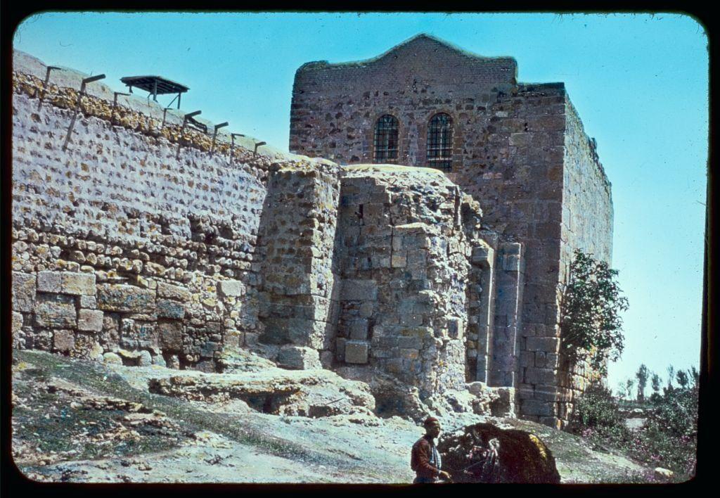 The image depicts a historical ruin with significant architectural features, possibly dating back to the Roman or Byzantine era. The structure is made of stone and brickwork, displaying signs of erosion and decay over time. It has two visible arched windows at its upper level and appears to have remnants of walls that once formed part of a larger building complex.
In front of this ancient edifice stands an individual wearing traditional clothing, engaging with what seems like livestock—a cow or bull—on the ground. The setting is outdoors under clear skies, suggesting a warm climate. Various elements such as vegetation around and scattered stones on the ground provide context to the environment in which these remnants are situated.
The image conveys a sense of historical exploration and preservation, capturing the essence of antiquity juxtaposed with contemporary human presence.