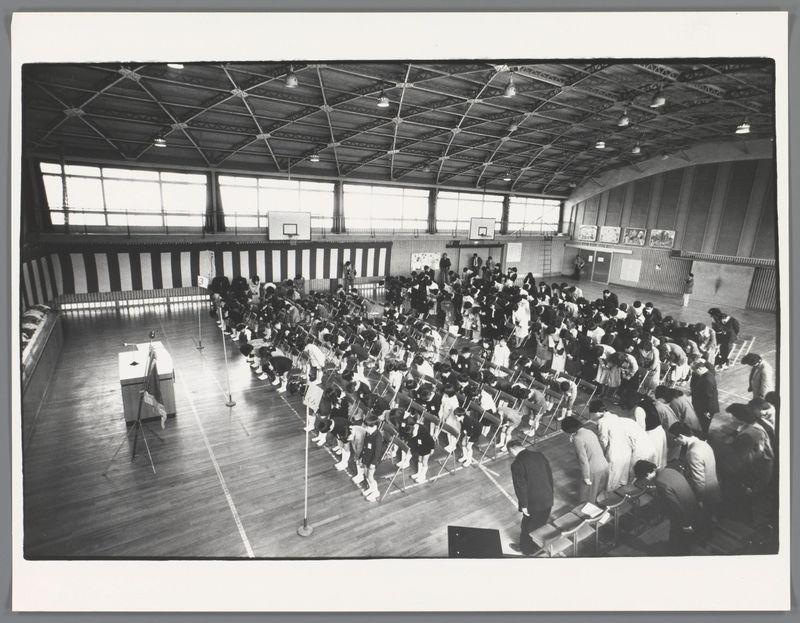 The image captures a lively indoor scene of what appears to be an educational or community gathering, possibly in Japan given the context provided. A large group of individuals is arranged on a gymnasium floor with wooden planks and white markings for sports lines. They are seated at desks facing towards one side where there seems to be some activity happening out of view; perhaps a teacher addressing them.

The setting features an expansive ceiling structure with trusses, indicative of the interior architecture typical in public or institutional buildings from mid-20th century Japan, possibly schools given the context. Natural light streams through windows at various heights along the walls, illuminating part of the room but leaving other areas in shadow, adding a dynamic contrast to the scene.

The group is dressed semi-formally; men wear suits and women are mostly seen wearing coats or blouses with skirts or slacks. A few individuals stand out due to their attire suggesting roles such as teachers or organizers. Several rows have children at desks who appear attentive but not engaged in writing, indicating a break possibly between sessions.

Various objects like whiteboards on the back wall suggest an educational environment, and banners above add decoration which might signify cultural events associated with this gathering. The atmosphere is one of orderly activity und [...]