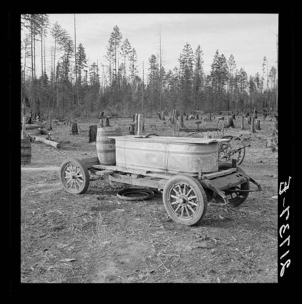 This black and white photo shows a wooden cart with two wheels, containing an old metal tub on the back. The tub appears to have been used for water storage or transportation in rural areas of Idaho. It is situated in what looks like a forest clearing with numerous trees cut down around it, possibly indicating logging activities. There are barrels nearby and remnants of other wood structures scattered throughout the area. The cart has visible wear marks on its wooden components, suggesting considerable use over time.