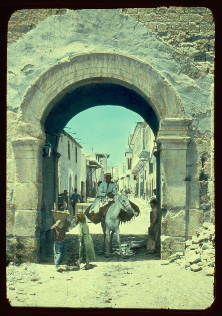 The image depicts a vibrant street scene through an arched gateway in what appears to be Damascus, Syria. The archway is weathered and constructed from large stone blocks with visible signs of aging or erosion.

Through the gate, we see a bustling alley lined by narrow buildings that exhibit traditional Middle Eastern architectural features such as tiled roofs and ornate facades. Several individuals are captured within this scene: an older man on horseback in the foreground wearing period-appropriate clothing, including a long coat, hat, and boots; two children near him, one carrying what seems like produce or goods.

The street is littered with debris, indicating perhaps a lack of urban sanitation at that time. The overall ambiance suggests a bygone era before modern infrastructure significantly altered such areas in historic cities.