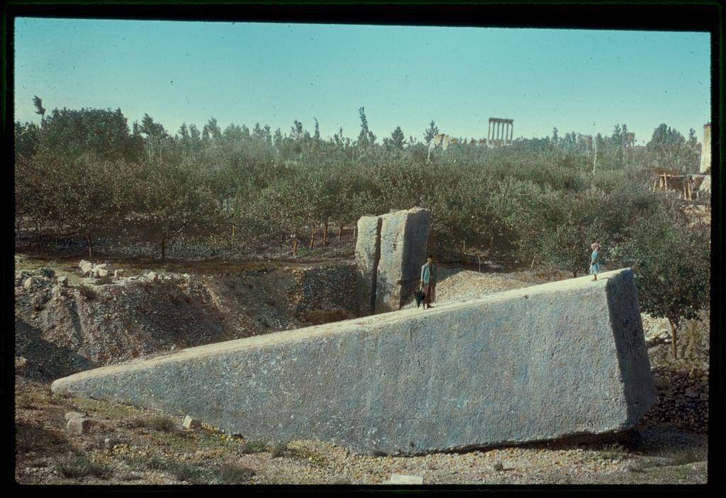 The image depicts a scene of ancient ruins and archaeological remains. In the foreground, there is a large stone structure that appears to be part of an old construction or fortification. The texture suggests it has been weathered by time.

On top of this massive stone platform, two individuals can be seen walking across its surface. They are dressed in what seems like traditional garments, indicating cultural attire possibly from the region where these ruins reside.

In the background, a landscape with trees and shrubs is visible under a clear sky. Amongst them stands another ancient structure that resembles columns or pillars, which could potentially belong to an archaeological site such as temples or palaces. The overall atmosphere of the image conveys historical significance and provides insight into past civilizations' architectural prowess.

The color tone has a slightly vintage feel with faded hues, likely due to aging effects on the photo itself. Additionally, some text is visible in the upper part of the photograph which may provide contextual information about its origins or history.