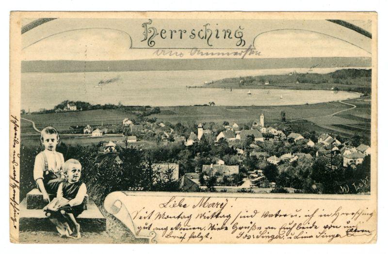 The image is a vintage black and white postcard featuring the word "Hersching" prominently at the top, suggesting it may be related to Herrsching am Ammersee in Germany. Below this title, there's an ornamental design that appears handwritten or stylized as if done with brush strokes. A scenic view of what looks like a lakeside village is depicted on the right side of the card, with houses and trees visible near water bodies under a partly cloudy sky.

In the foreground on the left side, two children are seated beside each other; one standing while holding something small in their hand, possibly food or an object. They seem to be posed for this photograph, as they look directly at the camera against a backdrop of what appears to be a stone ledge or wall.

At the bottom center of the image is cursive handwriting that reads "Like Memory," followed by more illegible script likely referring to personal notes from someone's perspective in memory. There are also multiple signatures and additional text, which due to their size and angle on the card, appear challenging to decipher clearly but include dates such as '20-74' or possibly something close.

The image seems historical with signs of wear typical for aged photographs, like creases along edges indicating it may have been folded. A handwritten annotation at the top left corner suggests that this was once owned by someone nam [...]