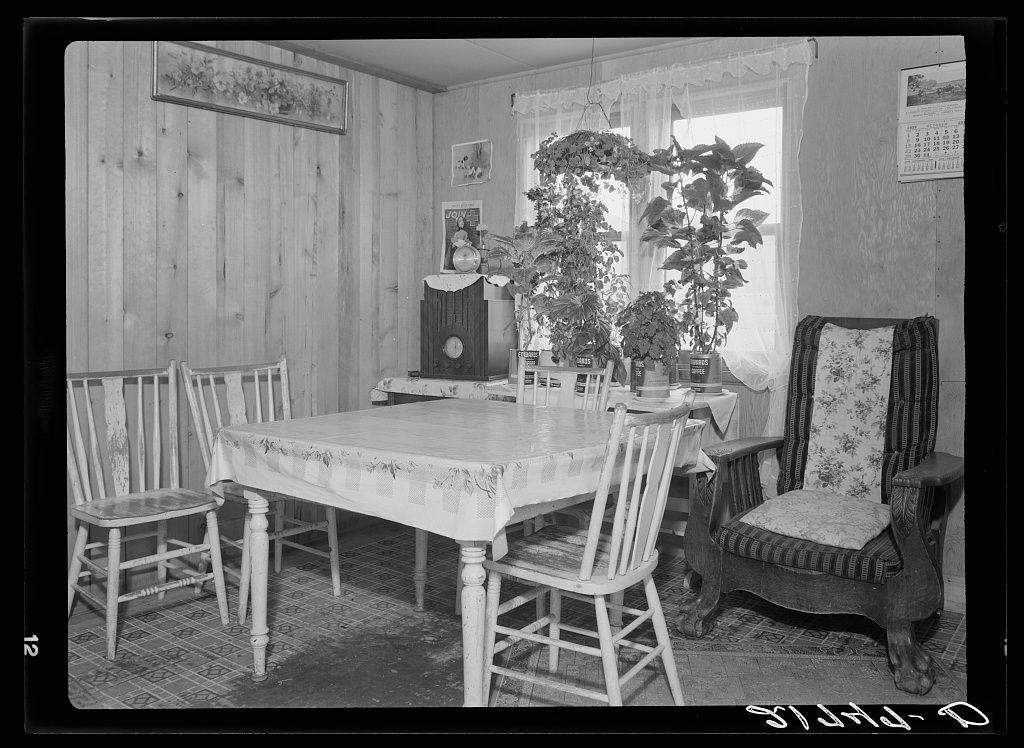 The image depicts a black and white photograph of an interior space, likely from the early to mid-20th century judging by its style. It shows a modest dining area within what appears to be a rural homestead in Idaho.

In this room, there is a wooden table with four chairs around it; one chair has ornate detailing while the others are plain and simple white-painted wood without decorative elements. The flooring features patterned linoleum tiles that have faded over time due to usage or aging. On the right side of the image stands an elegant striped armchair, suggesting a place for reading or contemplation.

A large potted plant with lush green leaves is situated near one window on the far left wall, adding a touch of life and color to the otherwise monochromatic scene. The wooden walls have character marks like knots which suggest natural timber construction typical in older homes.

Two windows can be seen; both are dressed with white curtains that allow some light into the room but also provide privacy when closed. A calender hangs on one wall, indicating a domestic setting where time and activities were tracked for convenience.

There's an old-fashioned radio console beside which several jars presumably containing food items or preserves suggest self-sufficiency typical of rural life at that period in America. The presence of houseplants could indicate the occupants' inc [...]