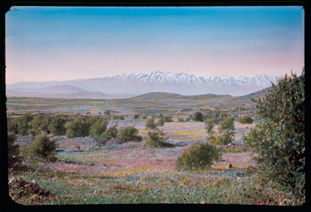 This image depicts a serene landscape featuring rolling hills with patches of green vegetation and wildflowers in vibrant shades of pink, purple, yellow, and red. In the distance, majestic snow-capped mountains form a striking backdrop against a clear blue sky tinged with soft pastel hues near the horizon. The overall scene exudes tranquility and natural beauty, likely captured during spring or early summer when flowers are blooming in such regions.
Additional information about this image suggests that it may be part of an archival collection from the 1950s to 1970s by Eric and Edith Matson. It is identified as Mt. Hermon viewed from the east, with a biblical reference to Luke 9:28 indicating its location in the vicinity of Damascus, Palmyra, and Baalbek. The image's dimensions are noted as 2 x 2 inches on one slide, emphasizing its small size within this collection.
