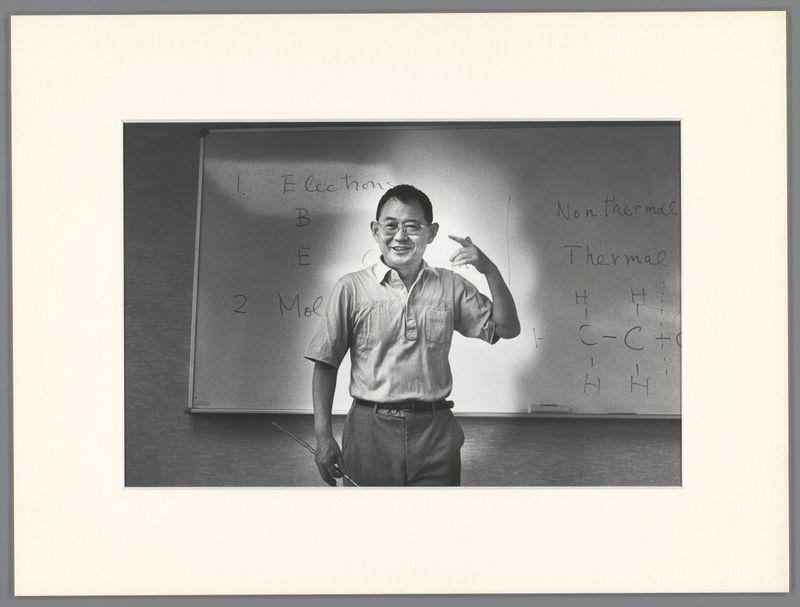A black and white photograph features a man standing in front of an educational chart. The individual appears to be gesturing towards the board, possibly explaining or teaching on electrical chemistry topics such as "Electrons," "Non-Thermal," "Thermal," "B E C Mo Lars" (likely meaning Boronborate Oxide), and various chemical notations like H-C+O-H -H. The man is wearing glasses, a short-sleeved collared shirt with text that seems to be Japanese characters or transliteration of the same on his chest pocket area. He has dark hair, parted in the middle, and appears engaged and animated during what might be an instructional session.